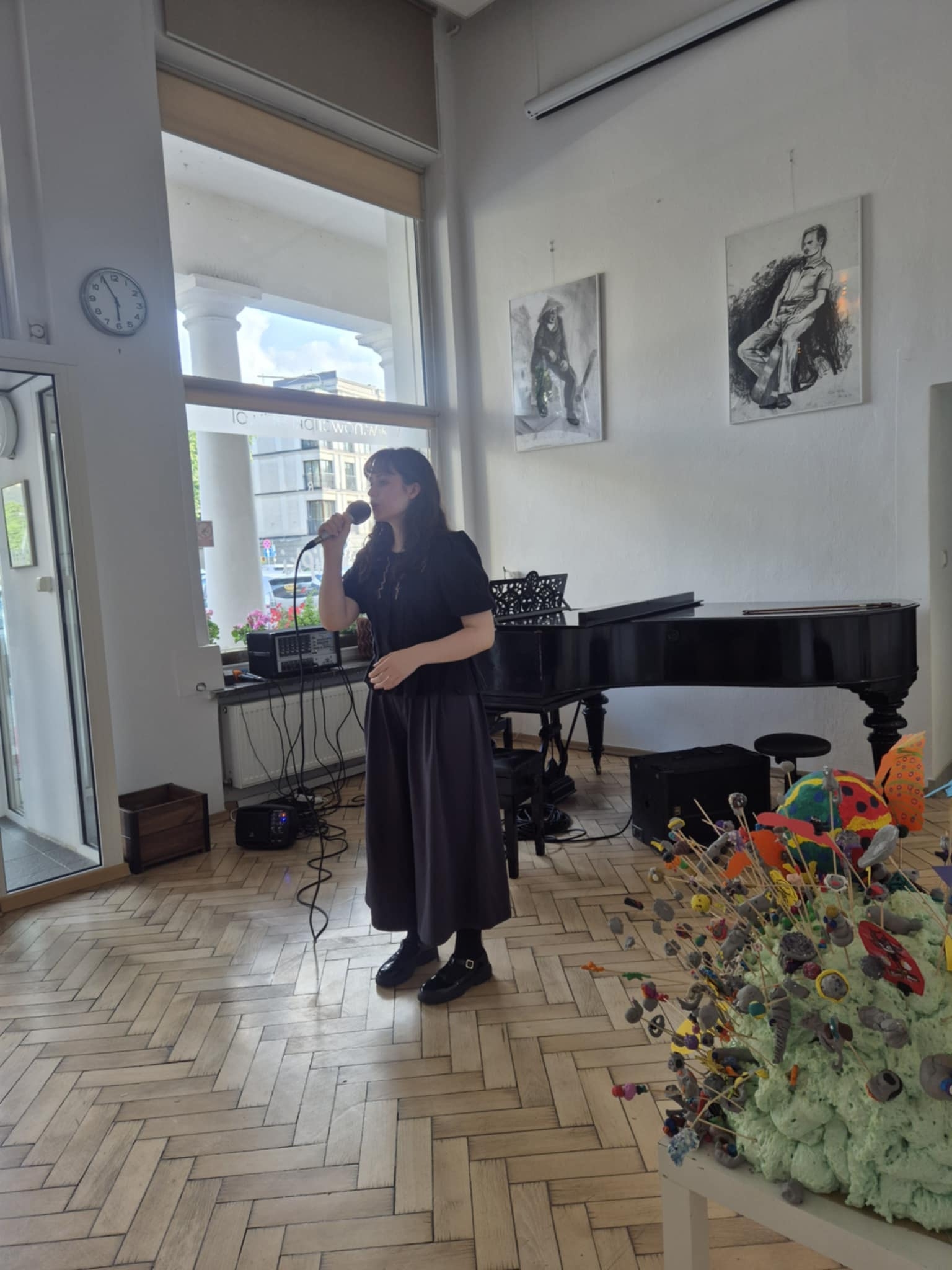 Solistka śpiewająca utwór z mikrofonem, w eleganckim stroju, z fortepianem w tle.